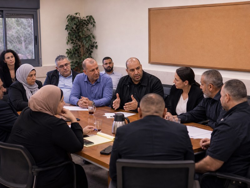מפגש קהילתי בין תושבים ערבים, אנשי משטרה ופקידי עירייה סביב שולחן ללא סמלים או טקסט קריא.