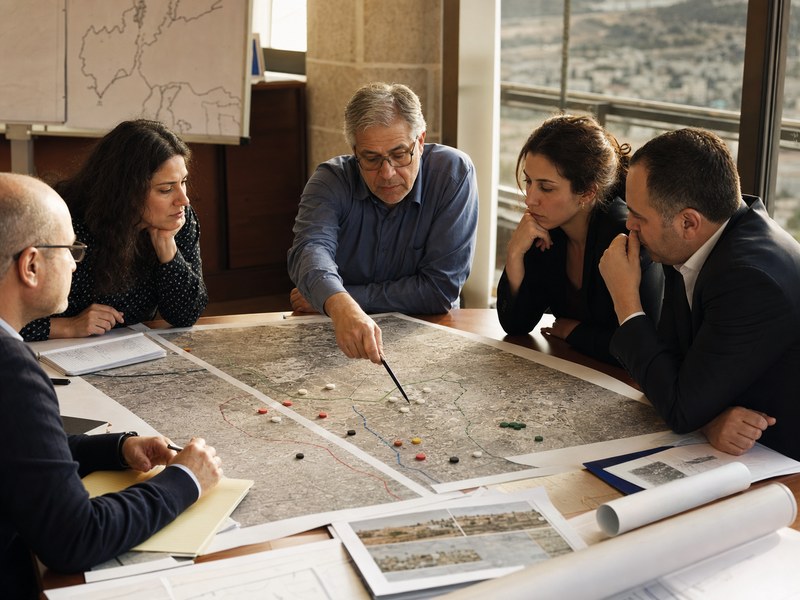יועצים אזרחיים ומשפטיים בוחנים מפות תכנון ריקות של תוואי ביטחוני סביב שולחן עבודה.