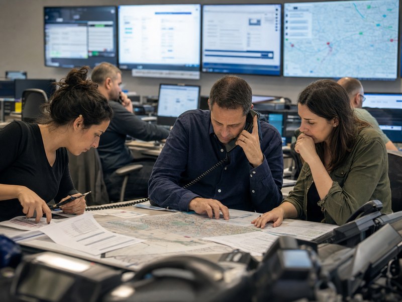 צוות תקשורת ציבורית בחדר מצב אזרחי בוחן מפות ומסכים מטושטשים בזמן משבר.