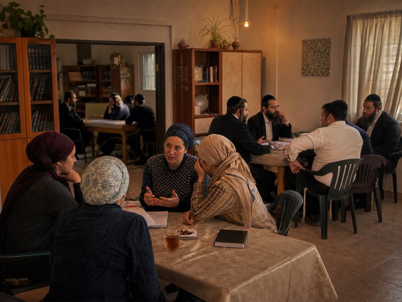 חדר קהילתי חרדי צנוע עם קבוצות שיחה ותה סביב שולחנות ללא טקסט קריא או סמלים רשמיים.